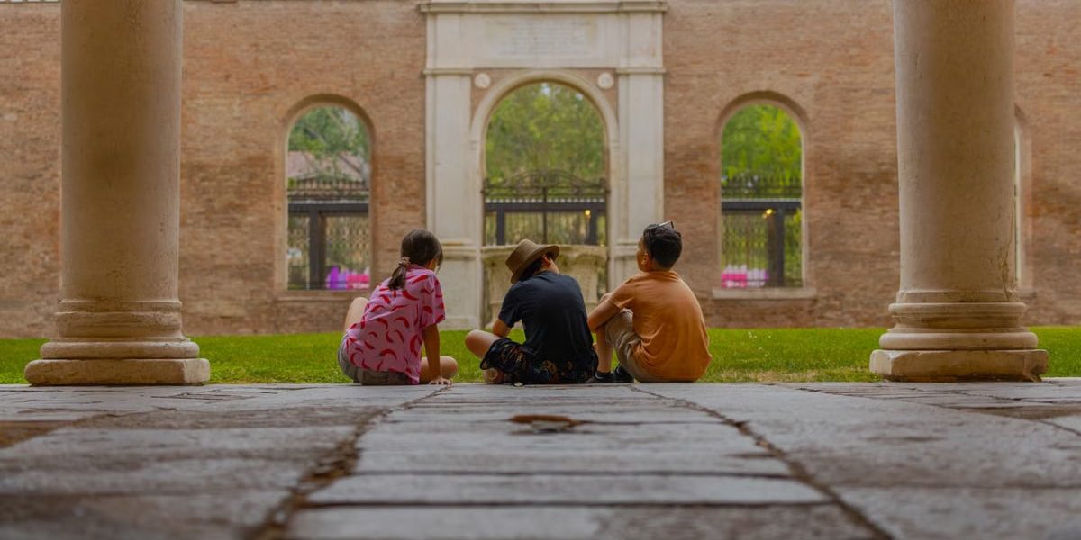 Three kids sit together in an ancient cloister, surrounded by marble pillars and lush greenery.