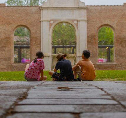 Three kids sit together in an ancient cloister, surrounded by marble pillars and lush greenery.